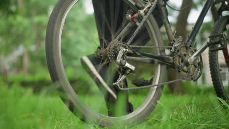 close-up of individual wearing black trousers and sneakers standing next to parked bicycle in grassy field, lifting rear tire and pedaling briefly, causing the tire to rotate with visible motion blur