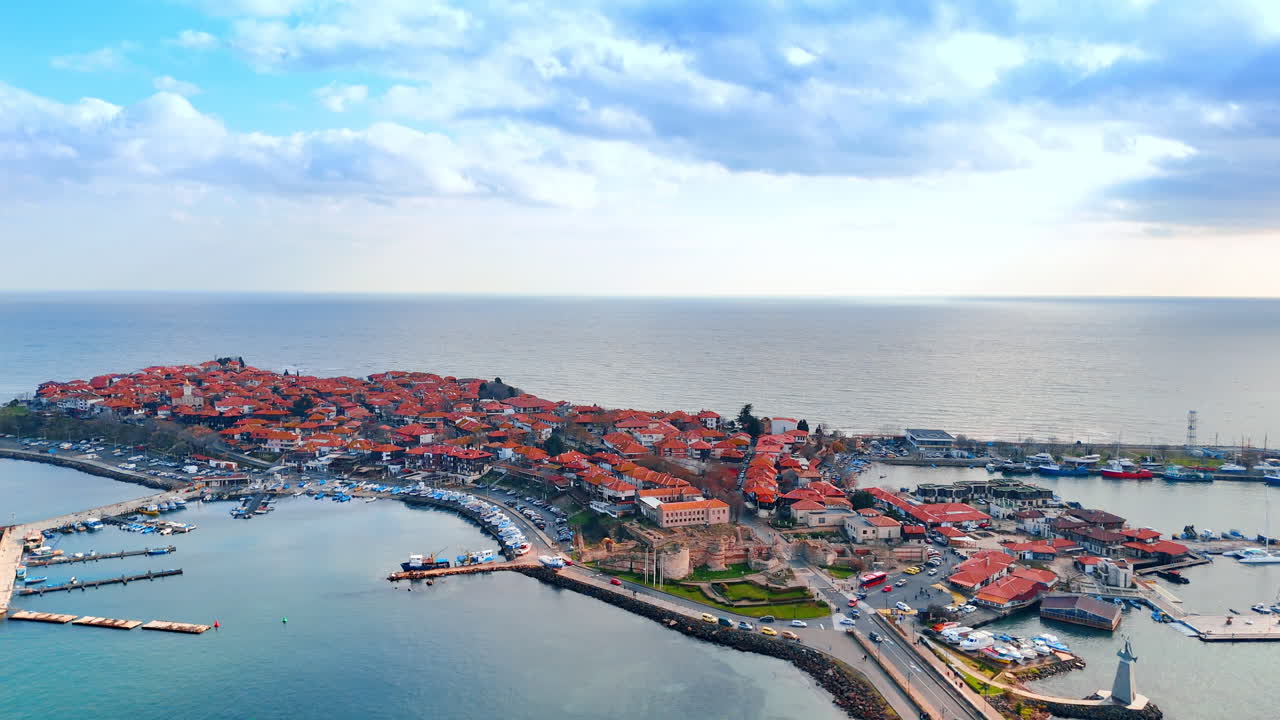 Cittadina costiera in riva al mare, illuminata da una luce calda. Una cittadina con tetti rossi si estende lungo la costa vicino all'acqua. Le barche sono ormeggiate al porto turistico. Il cielo è nuvoloso.