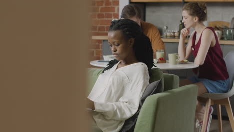 Chica-Trabajando-En-Una-Computadora-Portátil-Sentada-En-Un-Sofá-Mientras-Sus-Dos-Compañeros-De-Cuarto-Hablan-Juntos-Sentados-En-La-Mesa-De-La-Cocina