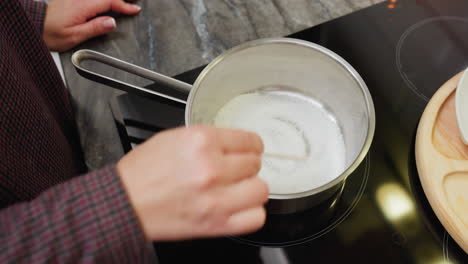 primer plano de una persona mezclando azúcar en una olla con una cuchara de madera, acción de cocción en la cocina, preparación de ingredientes para caramelo o postre en la estufa, azúcar en la sartén calentándose