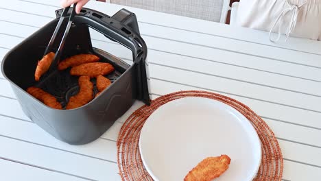 chicken tenders are removed from an air fryer and placed onto a plate in a bright kitchen setting