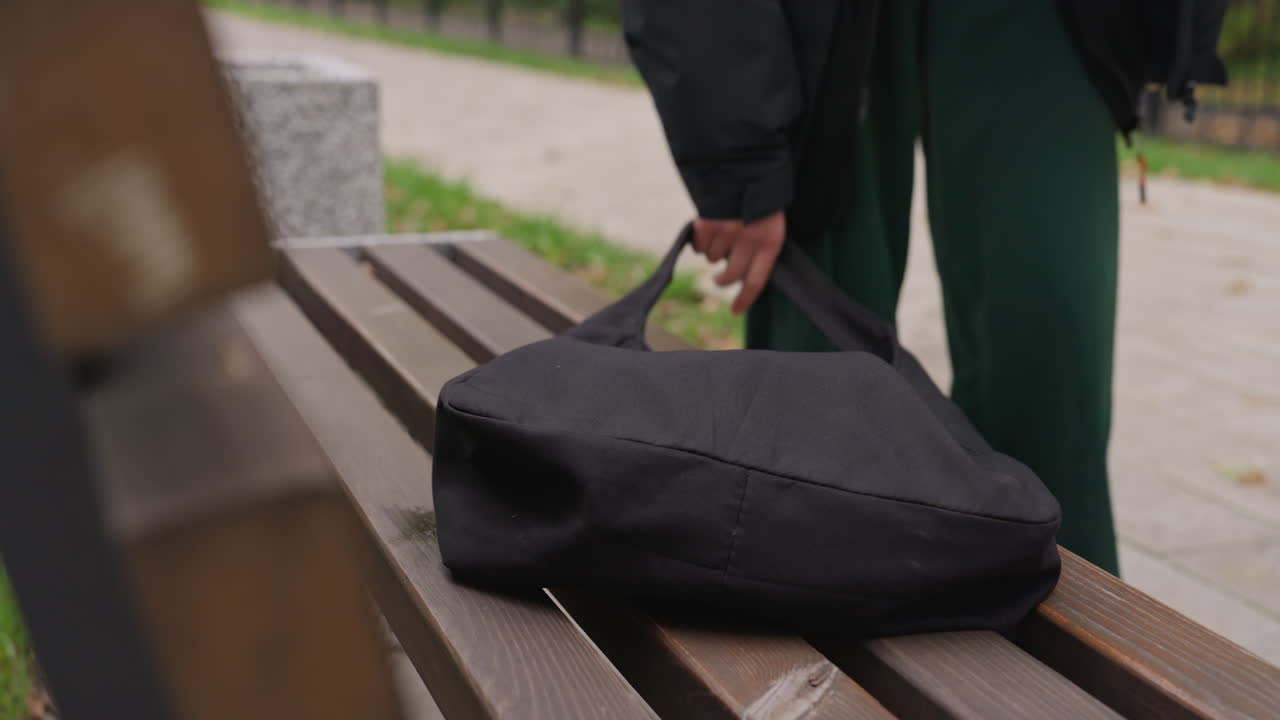 Una borsa nera su una panchina del parco, una persona se ne va. Una persona con pantaloni verdi appoggia una borsa e si allontana lungo un sentiero di ciottoli, listelli vuoti della panchina in primo piano, texture del legno umido, cielo nuvoloso che proietta una luce soffusa
