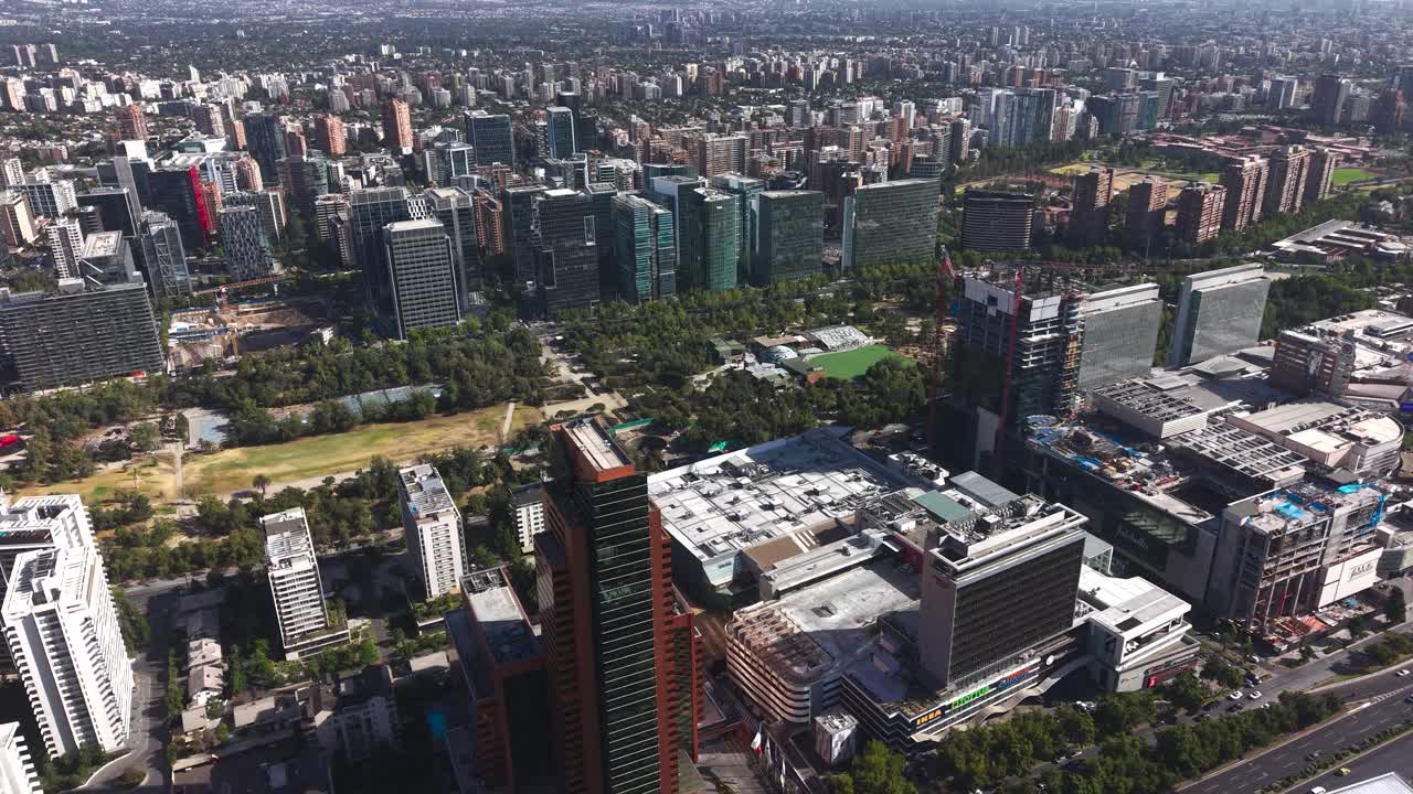 Filmagem aérea dinâmica circulando um arranha-céu moderno marrom-avermelhado em Las Condes, revelando os shopping centers, as torres de escritórios e o tecido urbano vibrante de Santiago, Chile