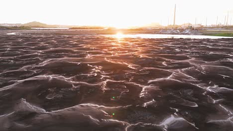 sunlight glistens against polluted water in asphalt lake at golden hour with oil refinery behind barren ground