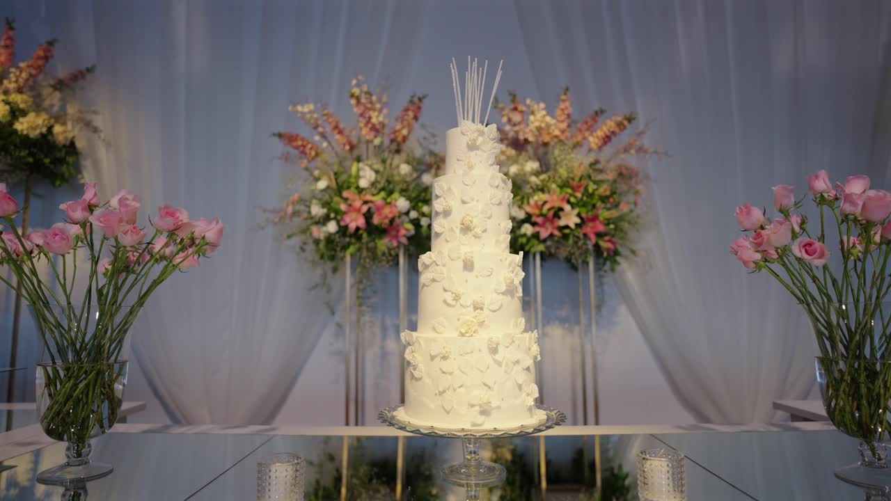 White multi tiered wedding cake reflecting on a glass table, showcasing elegance and celebration for a joyous occasion surrounded by beautiful floral decorations, slow motion orbit