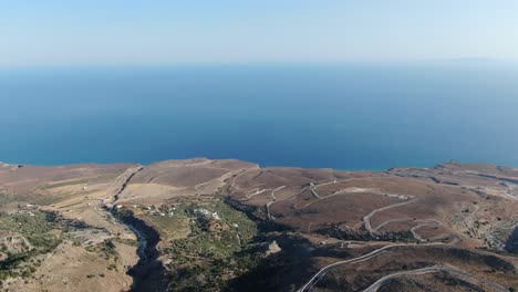 drone view in greece flying over a brown and green mountain with serpent road and sea on the horizon on a sunny day