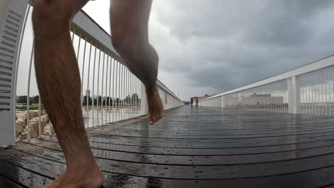 slow motion long hared man running without shoes over a foot bridge in the rain