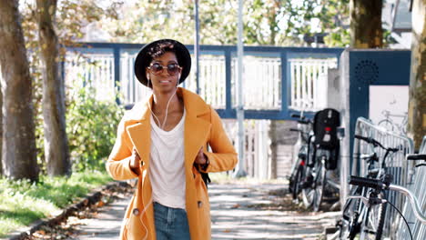 fashionable young black woman wearing a hat, sunglasses, an unbuttoned yellow pea coat and jeans, walking along a treelined city street listening to music