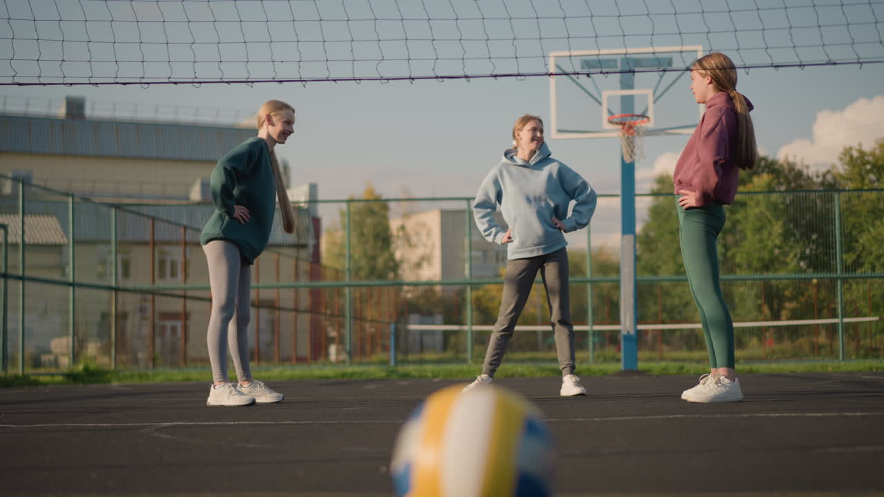 Tre kvinner i leggings og hettegensere deltar i en treningsaktivitet på en utendørs bane med en boligbygning og trær i bakgrunnen, som viser teamarbeid og bevegelse
