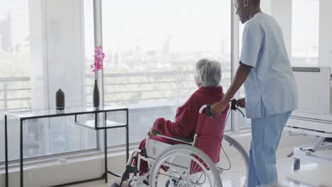 doctora diversa moviendo a una paciente anciana en silla de ruedas a la ventana de la sala, cámara lenta