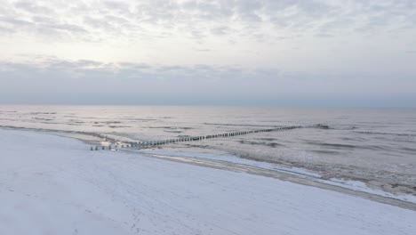 Aerial-establishing-view-of-an-old-wooden-pier-at-the-Baltic-sea-coastline,-overcast-winter-day,-white-sand-beach-covered-in-snow,-ice-on-wood-poles,-calm-seashore,-wide-drone-shot-moving-forward