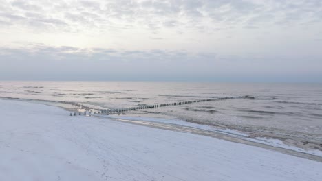 aerial establishing view of an old wooden pier at the baltic sea coastline, overcast winter day, white sand beach covered in snow, ice on wood poles, calm seashore, wide drone shot moving forward