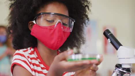niña con máscara facial y gafas protectoras sosteniendo un contenedor con productos químicos en el laboratorio