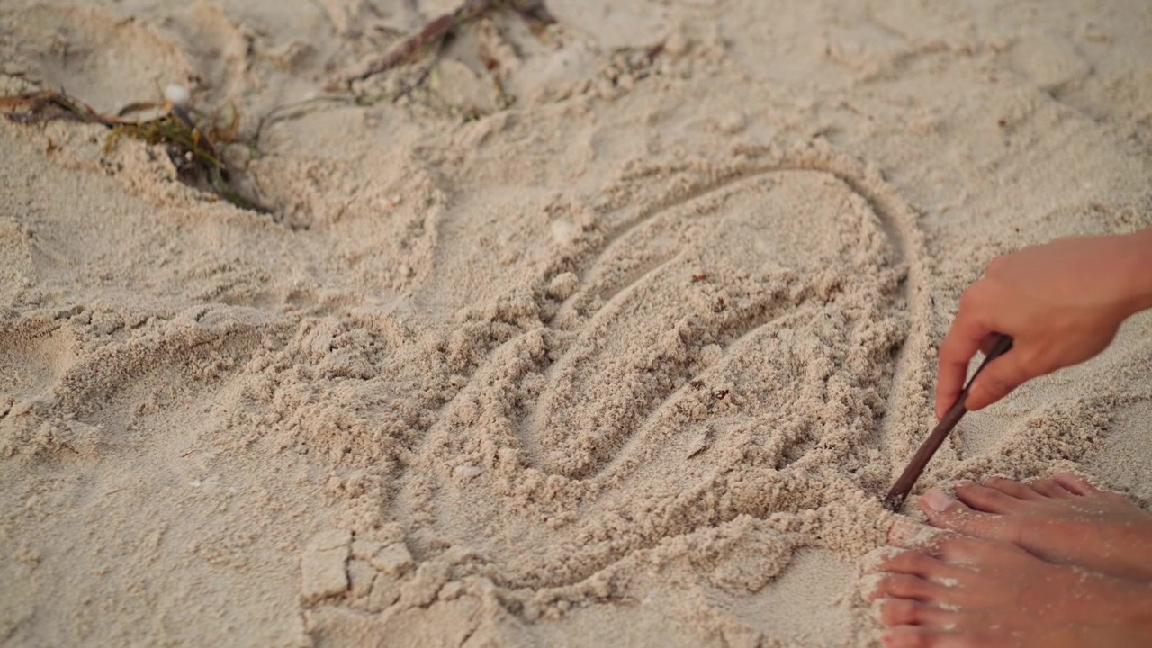 Händer ritar ett hjärta i strandsanden med en pinne bredvid fötterna, lekfullt romantiskt meddelande ristat i de texturerade sandkornen, spridd tång och fotspår vid kanten, varmt gyllene ljus och intim strandmiljö