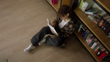 Top-view-of-a-girl-student-with-curly-hair-in-a-plaid-shirt-reading-a-book-near-shelves-with-books-in-the-library