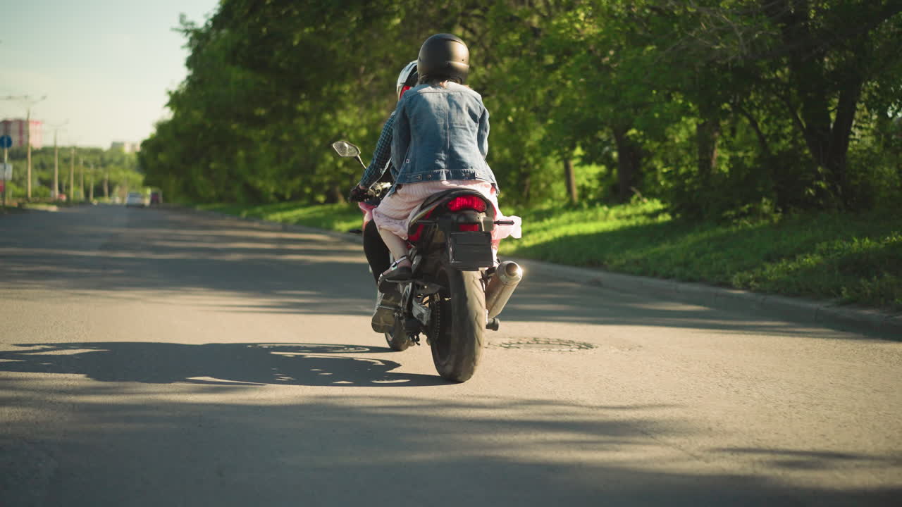 Two women riding a power bike, the passenger holding the rider tightly, as they travel on a shaded road lined with trees, the wind flutters the passenger s dress, with a distant car visible ahead