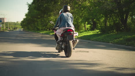 two women riding a power bike, the passenger holding the rider tightly, as they travel on a shaded road lined with trees, the wind flutters the passenger s dress, with a distant car visible ahead