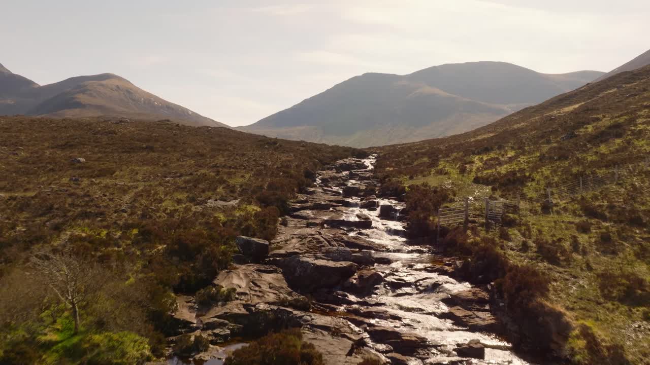 Aerial drone footage of a scenic river flowing through the rugged landscape of the Isle of Skye, featuring a beautiful waterfall surrounded by dramatic hills and untouched nature