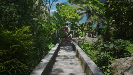 A-woman-carefully-making-her-way-down-a-staircase-set-within-a-tropical-paradise,-with-the-mesmerizing-and-endless-ocean-as-the-background