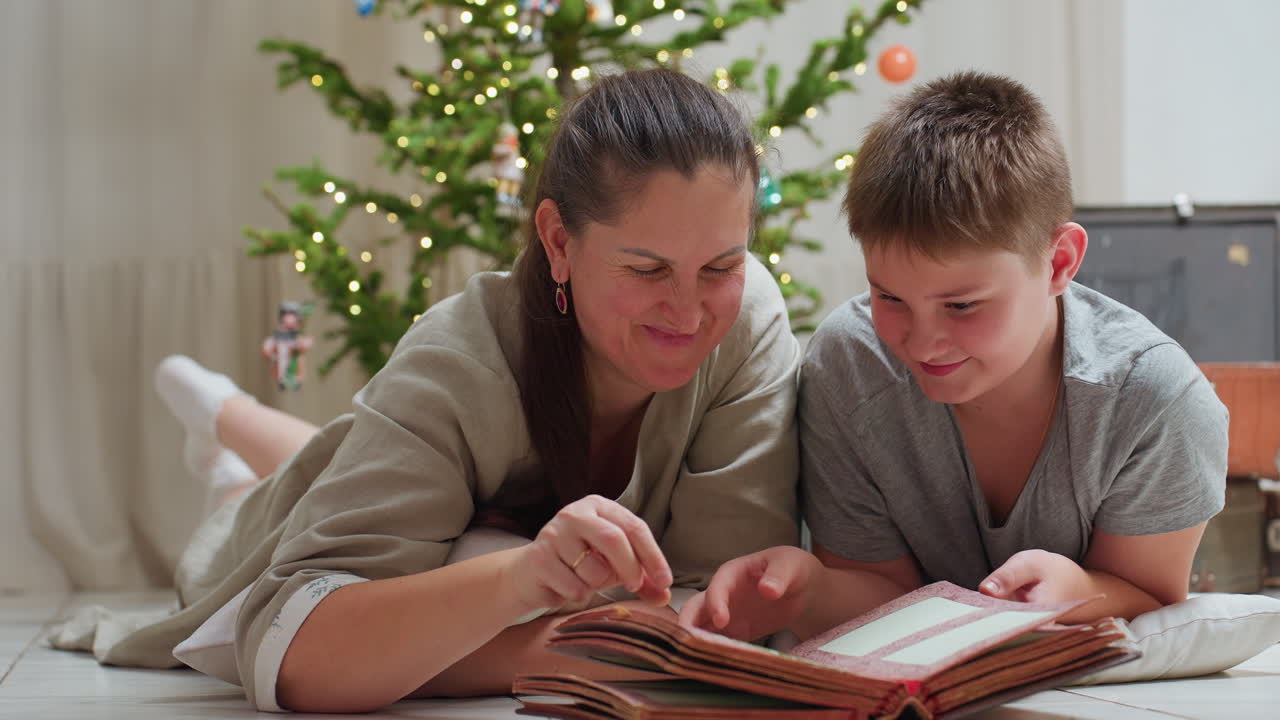 mother and son lying on marble floor smiling while opening vintage book cozy festive home setting background with decorated christmas tree soft light joyful bonding moment