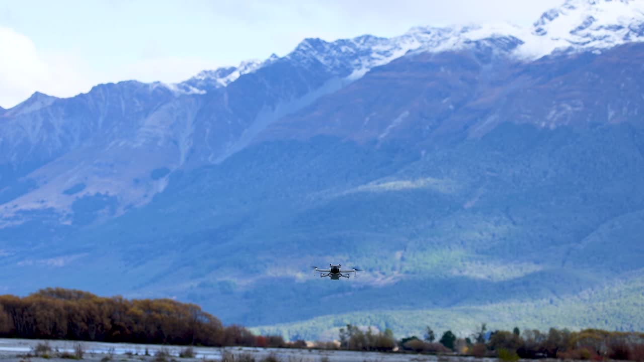 Quadcopter drone flies forward above river, framed by mountains, under bright natural daylight
