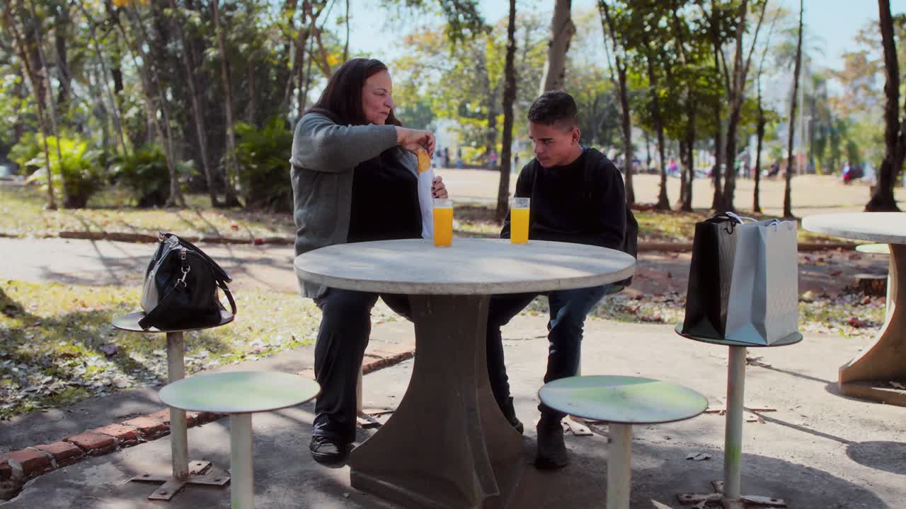 Woman and young man sharing a snack and drinks at an outdoor table in a park