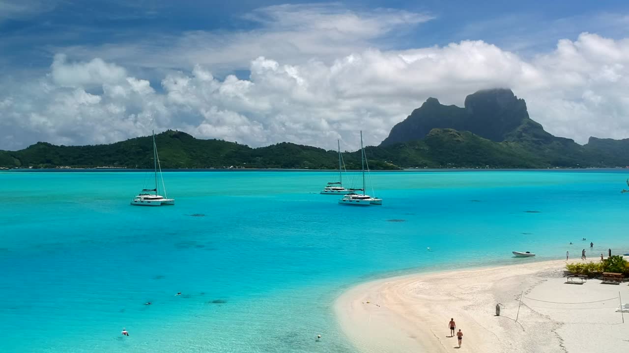 Drone shot of Bora Bora from a private Motu. perfect blue ocean with a few catamaran anchor in front of Bora Bora. People walk on the beach in french polynesia