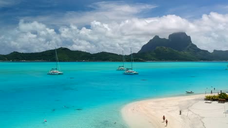 drone shot of bora bora from a private motu. perfect blue ocean with a few catamaran anchor in front of bora bora. people walk on the beach in french polynesia