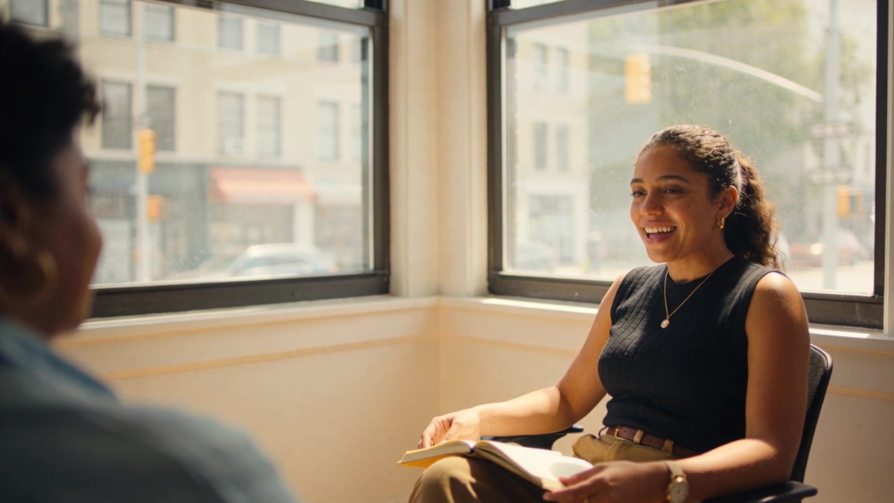 Talking woman in sleeveless black top responding to prompt in corner office, holding notebook