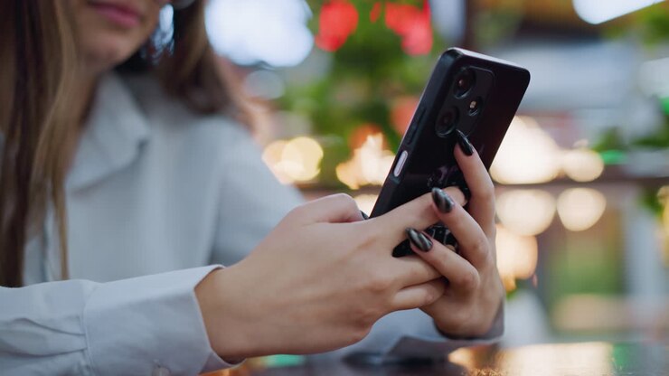 Primo piano di una donna con unghie nere laccate che chatta al telefono, con uno sfondo sfocato dai colori tenui e vivaci caratterizzato da luci bokeh, che mostra un momento di comunicazione moderno ed elegante in un ambiente raffinato