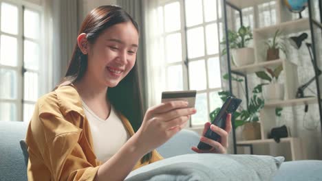 Happy Asian Woman Sitting On Couch In Living Room Holding Credit Card And Using Smartphone Shopping Online
