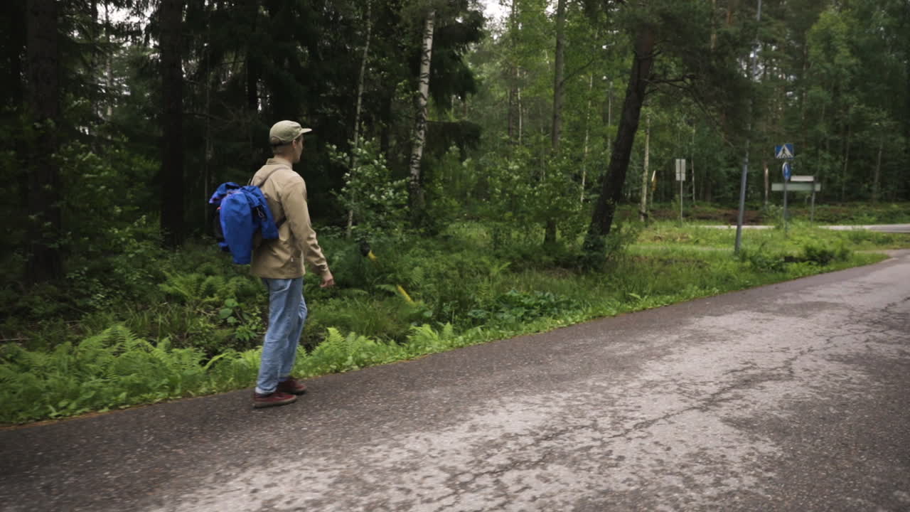 un joven con gorra y mochila camina al costado de la carretera por un bosque verde
