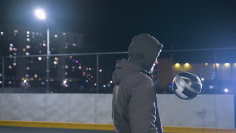 joven atleta entrenando por la noche jugando al fútbol hábilmente con la rodilla en el campo deportivo al aire libre bajo la luz de la calle brillante, contra edificios residenciales en un entorno urbano