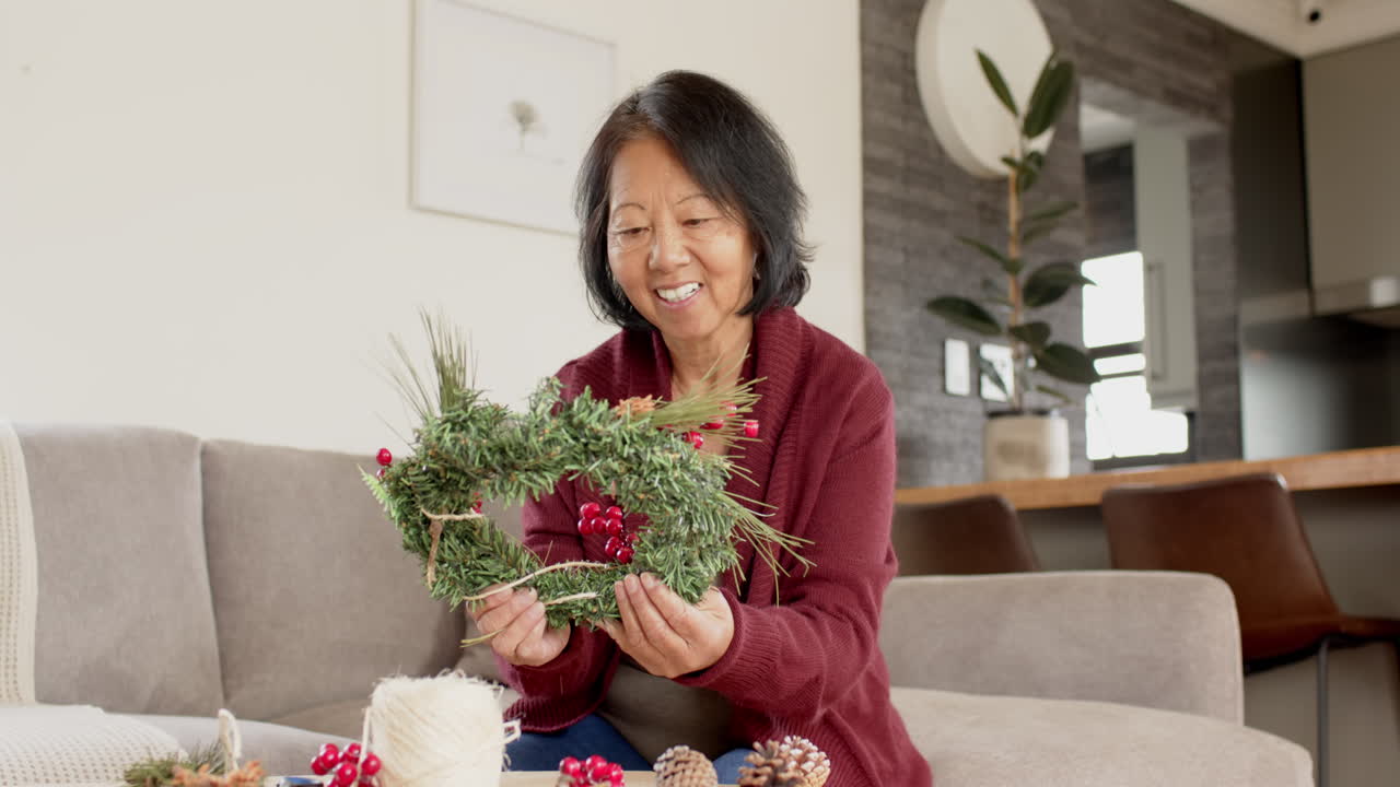 Smiling woman crafting Christmas wreath at home, enjoying festive creativity