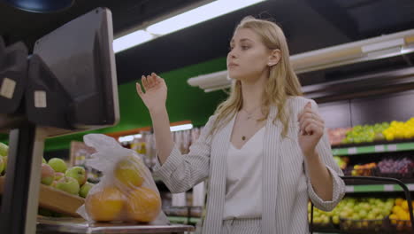 a blonde girl in a supermarket weighs oranges on an electronic scale pressing the display standing with a basket in her hands