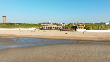 long aerial shot moving away from people enjoying their drinks on a beach club terrace outside a picturesque little coastal town, revealing the sea and a large, green rural area