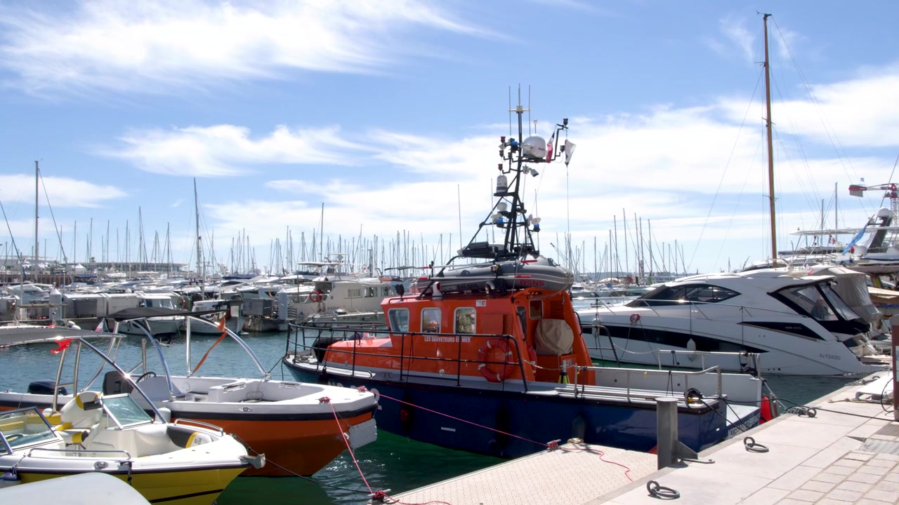 Un'imbarcazione di salvataggio arancione è ormeggiata tra yacht di lusso in un soleggiato porto della Costa Azzurra