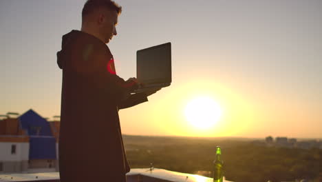 a male stockbroker freelancer stands on a rooftop at sunset with a laptop and types on a keyboard with his fingers looking at the cityscape from a bird's eye view.