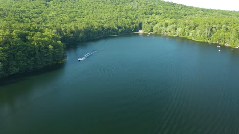 aerial drone shot flying high over a motor boat pulling a water skier along the lake shore on a sunny day in new hampshire, usa