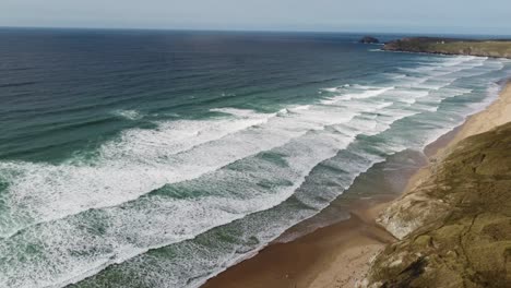 Eine-Drohnenaufnahme-Eines-Atemberaubenden-Strandes-In-Cornwall,-Der-Eine-Riesige-Länge-Goldenen-Sandes-Mit-An-Land-Kommenden-Surfwellen-Und-Einer-Wunderschönen-Landzunge-In-Der-Ferne-Enthüllt