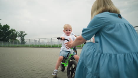 a woman stretches out her hand with a welcoming gesture as her younger son rides a bicycle happily towards her, while her older son follows closely behind on the same path