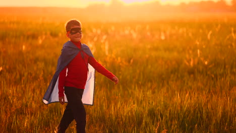 a boy dressed as a super hero standing in a mask and a red cloak runs laughing at the sunset in the summer field. summer night the boy dreams and heroic deeds and comic books