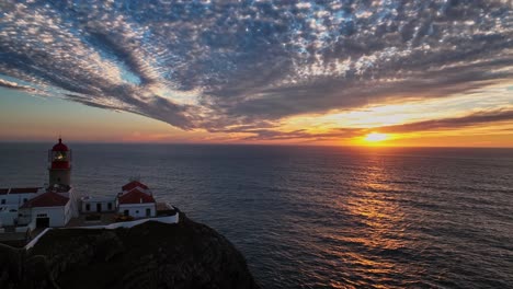a wider aerial view of the lighthouse and seagulls flying over the dramatic cliffs surrounding it at sunset.
