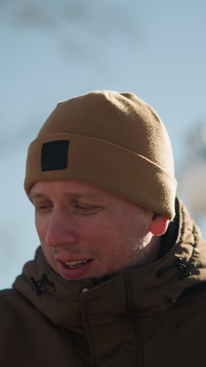un primer plano de un adulto con una chaqueta marrón y una gorra beige, hablando mientras camina al aire libre en un día brillante y soleado, con una vista borrosa de un árbol desnudo y un cielo despejado