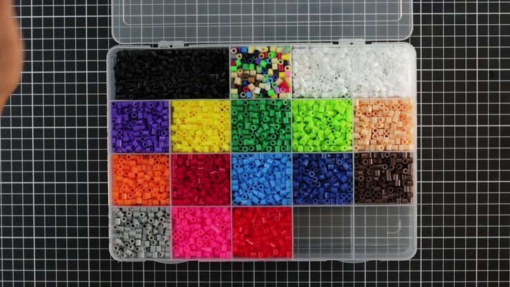 Overhead view of hands opening a case filled with colorful MIDI fusion beads on grid background