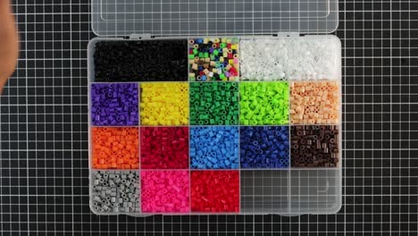 Overhead-view-of-hands-opening-a-case-filled-with-colorful-MIDI-fusion-beads-on-grid-background