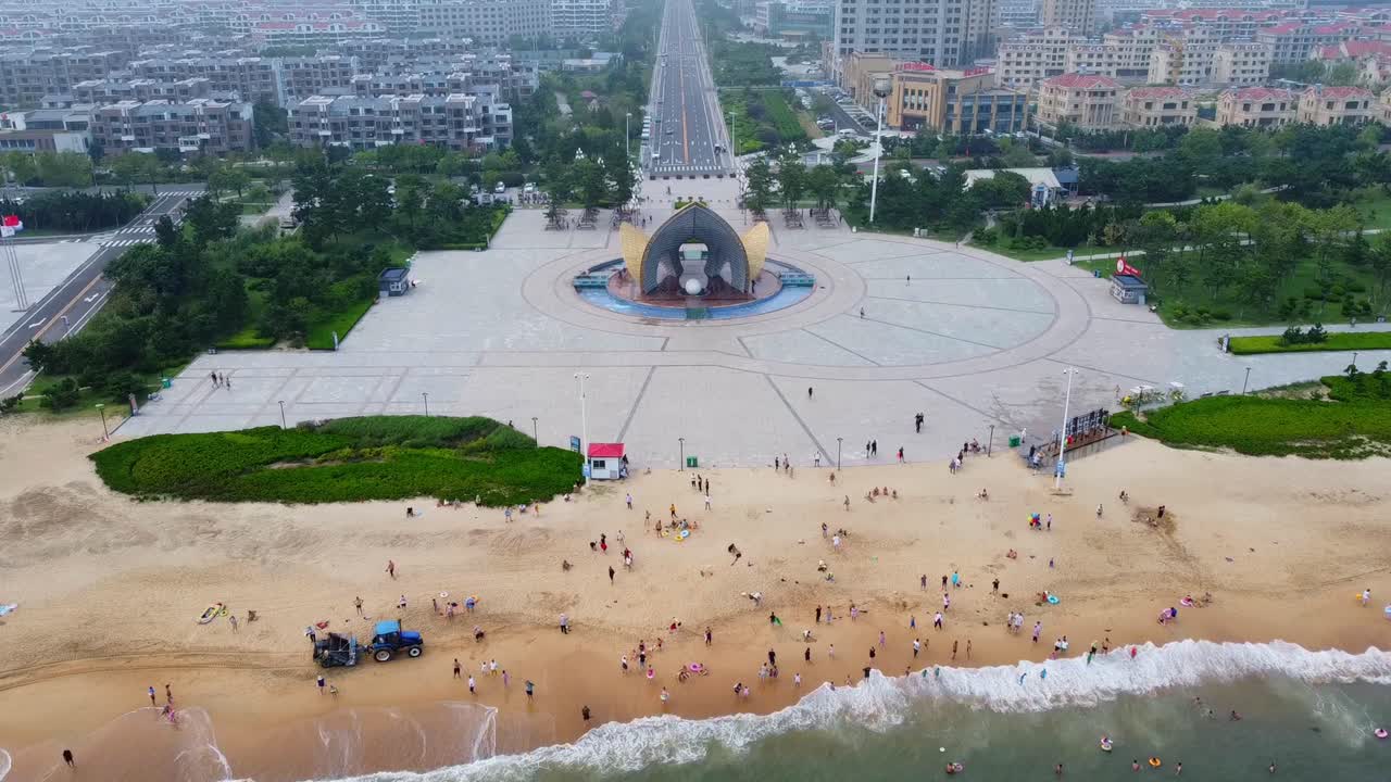 Crowded sandy beach with beautiful powerful waves crashing the shore and natural park, cityscape in background - high aerial view