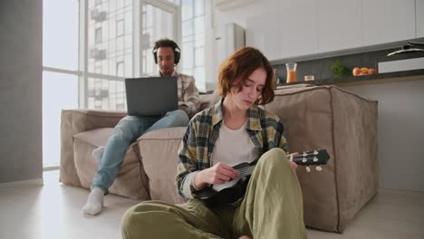 a young girl with a bob hairstyle in a green checkered shirt sits near the sofa on the floor and plays the ukulele while her boyfriend in modern black headphones works at a gray laptop in a modern studio apartment