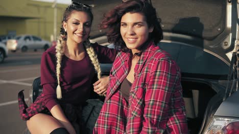 two young beautiful women sitting in a car trunk in the parking by the shopping mall during sunny day, smiling and looking in the camera