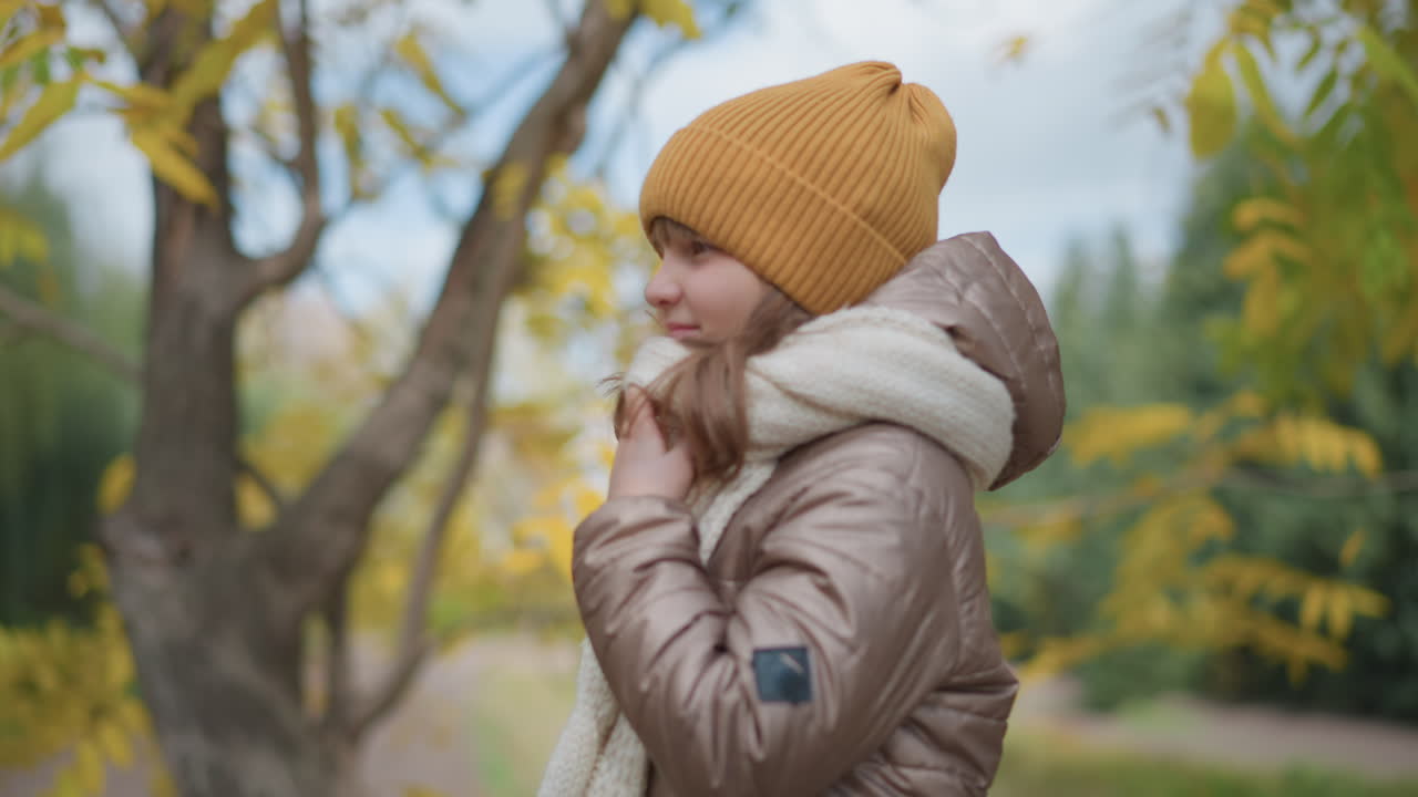 side view of cheerful girl wearing mustard beanie and puffy jacket smiling softly while gently holding cozy white scarf around neck in vibrant autumn park surrounded by golden and green foliage